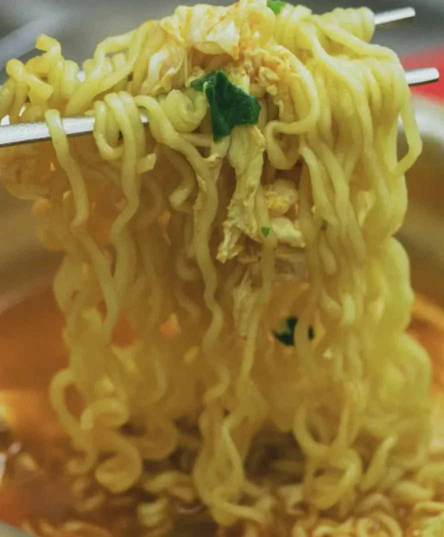 Fios de ramyeon com acelga e cebolinha erguidos com hashi de metal sobre a panela