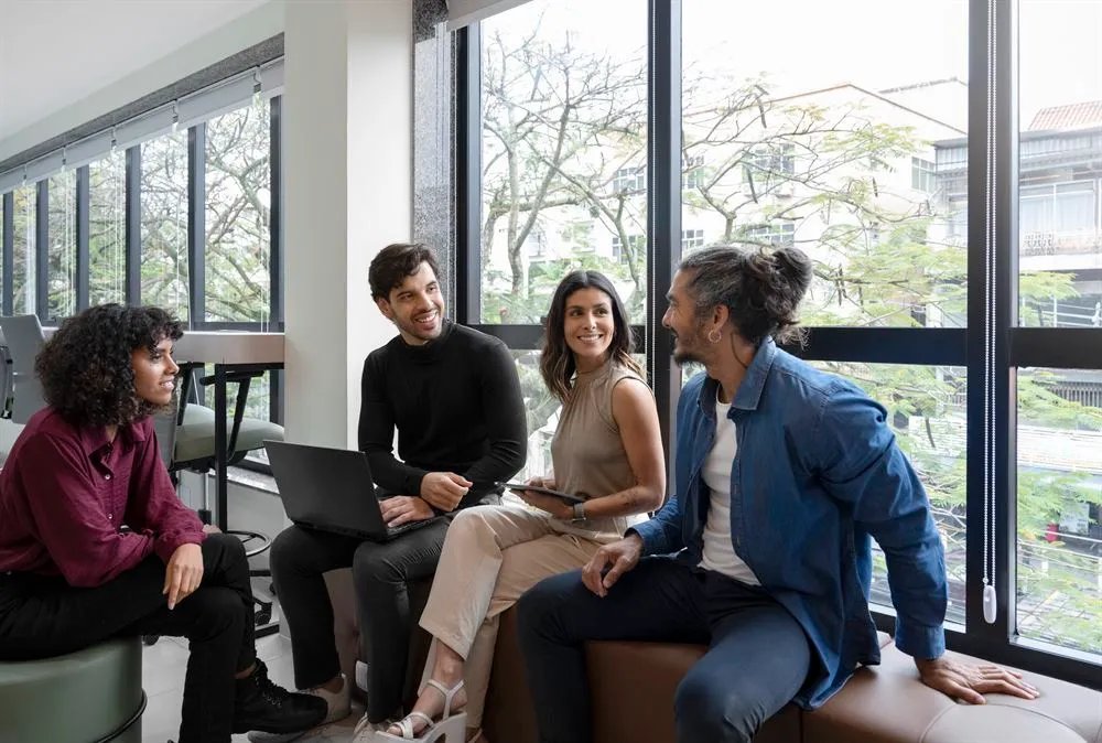 Quatro profissionais conversando com notebook e tablet em um escritório com janelas amplas