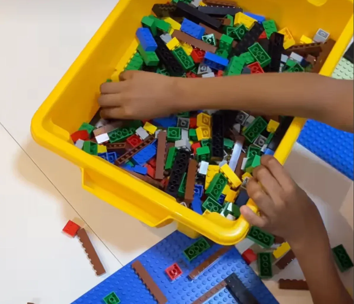Criança escolhe peças coloridas de LEGO dentro de uma caixa amarela, com uma base azul montada ao lado