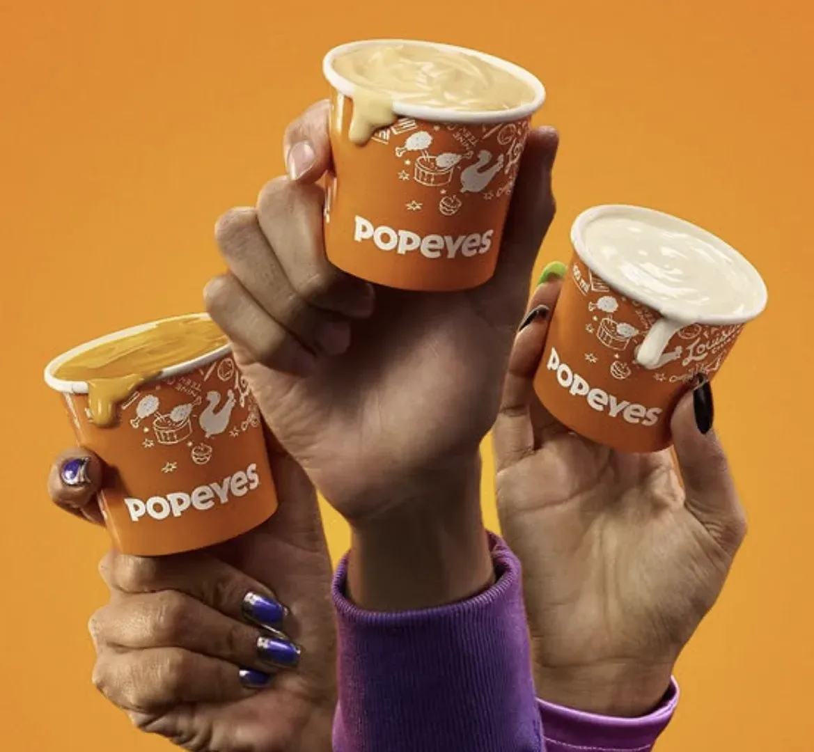 Cartaz da Batalha dos Queijos do Popeyes com os molhos cheddar, requeijão e provolone e os artistas Budah, Jotapê e Slipmami
