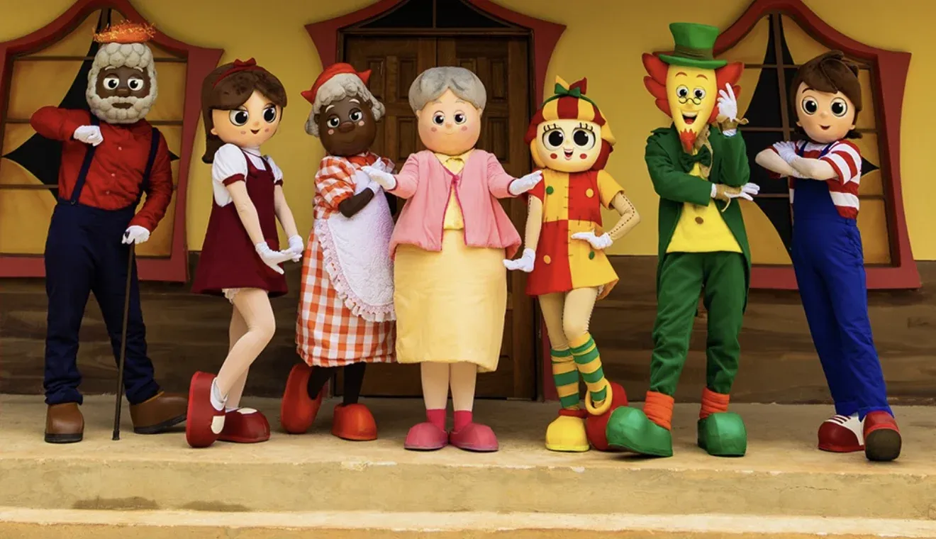 Emília, Narizinho, Pedrinho, Visconde, Dona Benta, Tia Nastácia e Tio Barnabé lado a lado na varanda do casarão