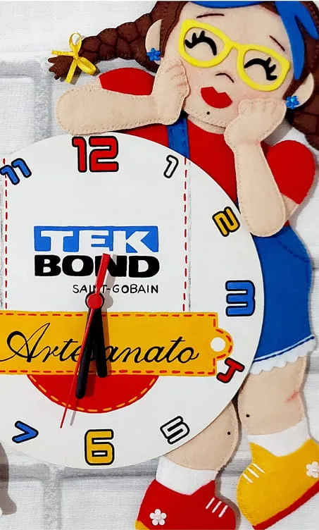Tekinha em feltro segurando o rosto, montada sobre um relógio de parede branco com o logo Tekbond Artesanato