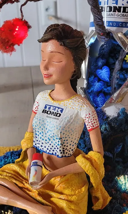 Boneca Tekinha esculpida em pose de meditação, com saia amarela e camiseta Tekbond, em cenário com árvore vermelha